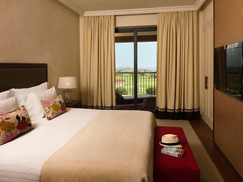 Schlafzimmer mit großem Bett, brauner Decke, zwei Kissen, roter Bank, TV und Blick auf Balkon mit Meer.