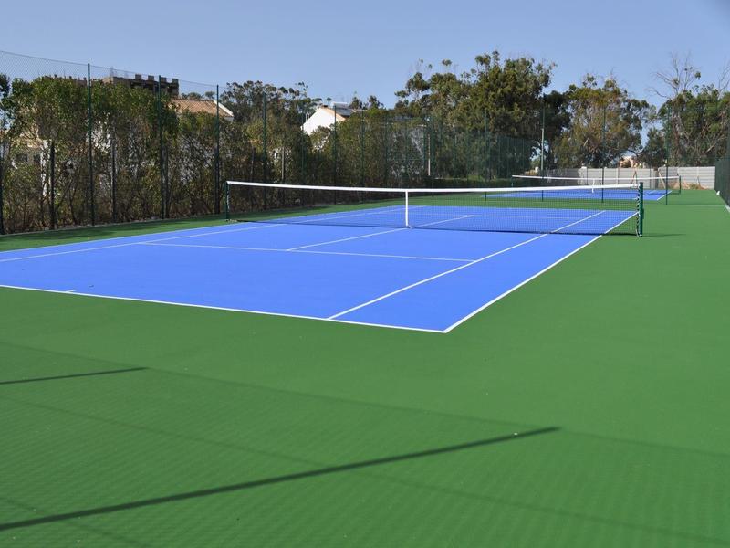 Freiluft-Tennisplatz mit blauem Spielfeld und grünem Umfeld unter klarem Himmel.