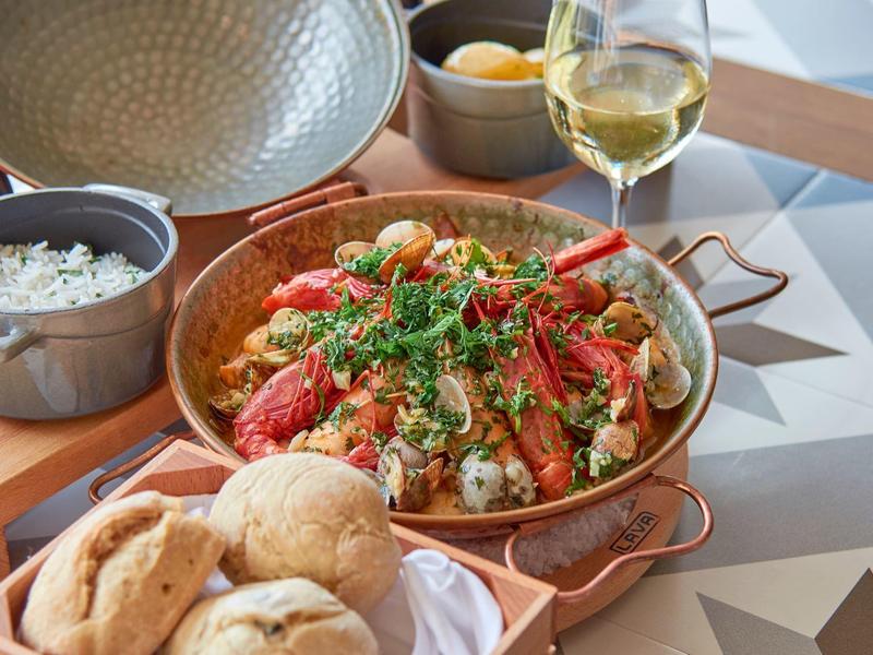 Tabakpfanne mit Hummer, Reis, Brot und Weißwein auf gemusterter Tischoberfläche serviert.