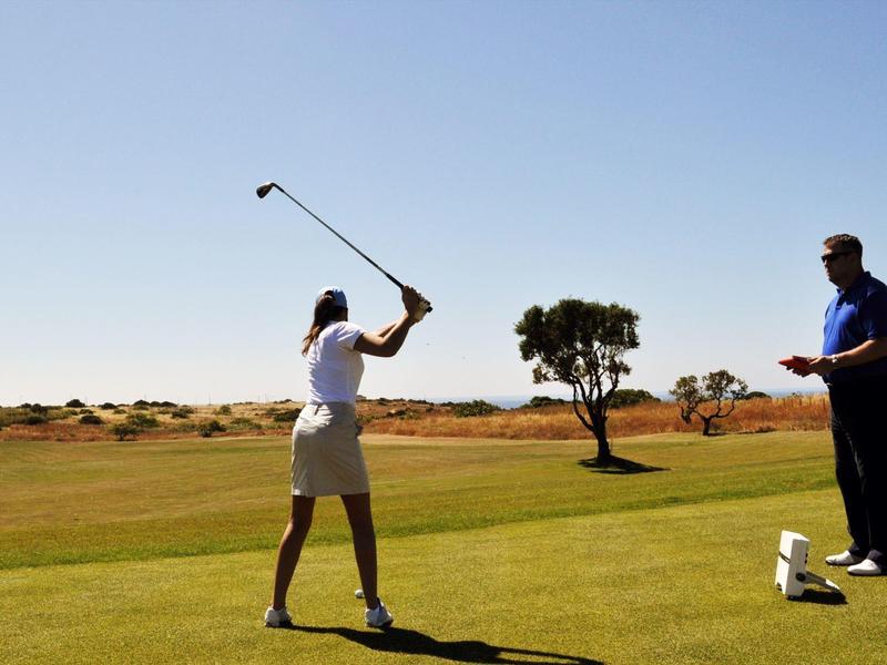 Zwei Personen auf einem Golfplatz, eine schlägt den Ball, die andere beobachtet.