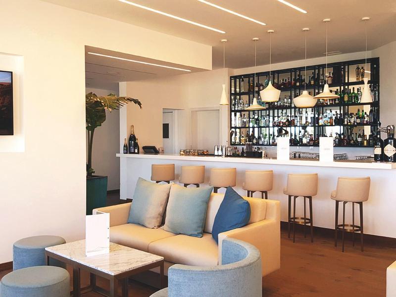 Modernes Cafè mit hellem Sofa, blauen Kissen, Barhockern und beleuchtetem Weinregal.