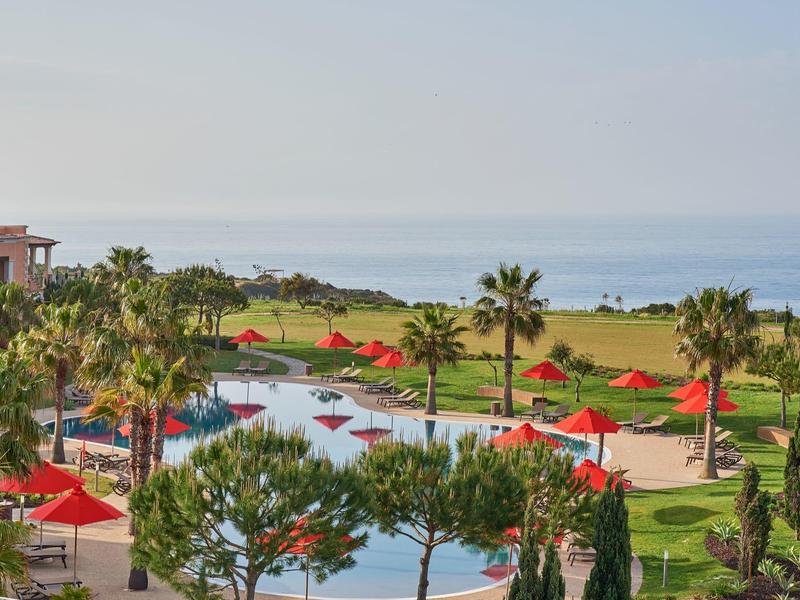 Großer Außenpool mit roten Sonnenschirmen, Palmen und Blick auf das Meer im Hintergrund.