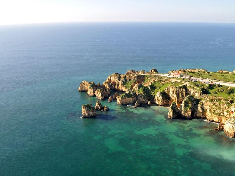 Felsige Klippen mit grün bewachsenem Gebiet neben klarem blauem Meer unter hellem Himmel.