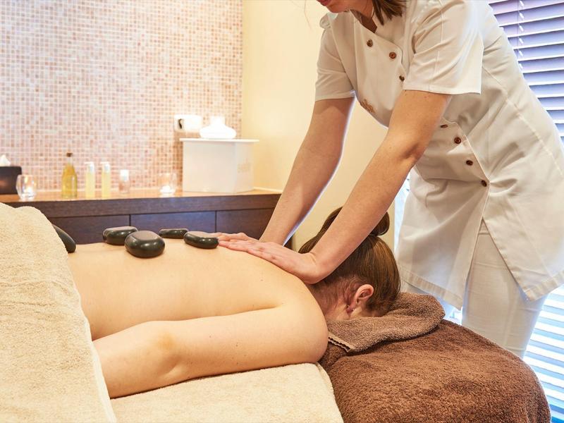 Frau liegt entspannt auf Massageliege, während Therapeutin Hot-Stone-Massage am Rücken macht.