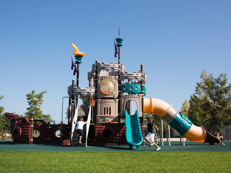 Spielplatz mit Piratenschiff-Klettergerüst, Folienrutschen und grüner Wiese unter blauem Himmel.