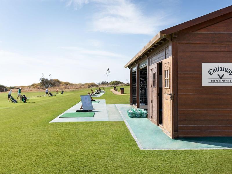 Grünes Golfplatzgelände mit Übungsbereich, braunem Holzhäuschen und blauem Himmel im Hintergrund.