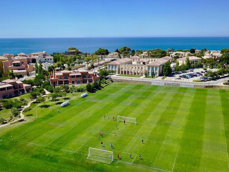 Großes grünes Fußballfeld mit Toren vor einer Küstenstadt und blauem Himmel.