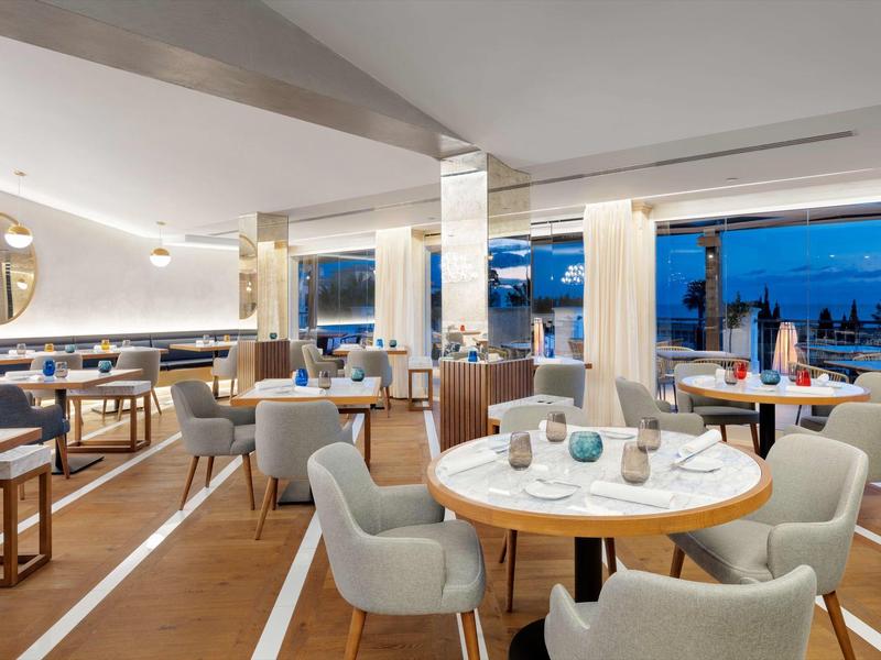 Moderner, heller Restaurantraum mit weißen Tischen, grauen Stühlen und großen Fenstern mit Ausblick.