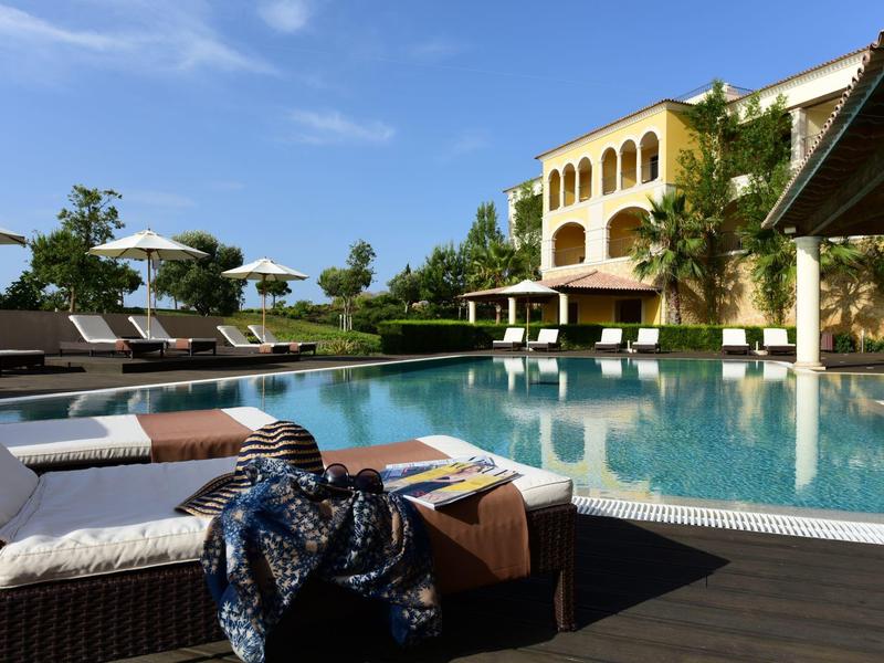 Luxuriöser Hotelpool mit Liegestühlen, Sonnenschirmen und einer dreistöckigen gelben Villa bei blauem Himmel.