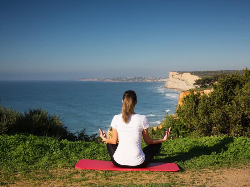 Frau sitzt auf roter Matte, meditiert am Meer mit Klippen und blauem Himmel im Hintergrund.
