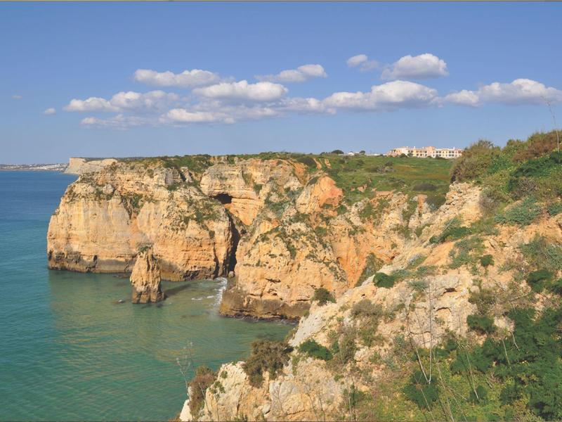 Klippen mit orange-braunem Felsen an ruhigem türkisfarbenem Meer unter blauem Himmel mit Wolken.