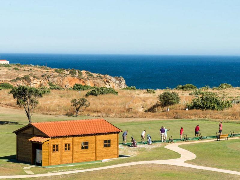 Grüne Golfanlage mit kleinem Holzhaus, Spielern, Meer im Hintergrund und klarem blauen Himmel.