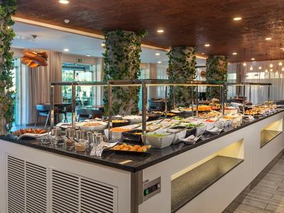 Buffet mit vielfältigen Speisen auf langer Theke, begrünt von Säulen mit Pflanzen, warme Innenbeleuchtung.