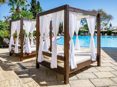 Holz-Überdachungen mit weißen Vorhängen stehen am Pool auf Steinfliesen unter blauem Himmel.
