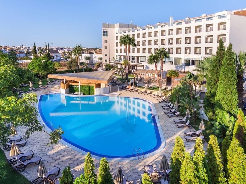 Großes Hotel mit rundem Pool, Liegestühlen, Palmen und grüner Umgebung unter blauem Himmel.