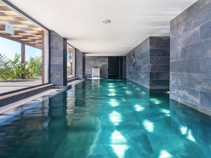 Innen beheizter Pool mit glatten grauen Wänden, großen Fenstern und Sonnenlichtreflexionen im Wasser.