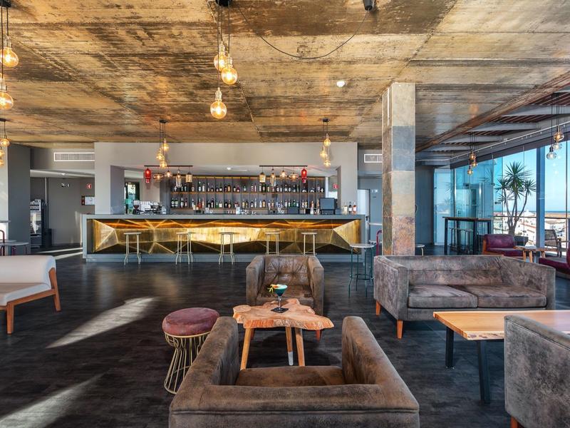 Moderner Barbereich mit Holztheke, hängenden Glühbirnen und gemütlichen Samtsofas in warmem Licht.