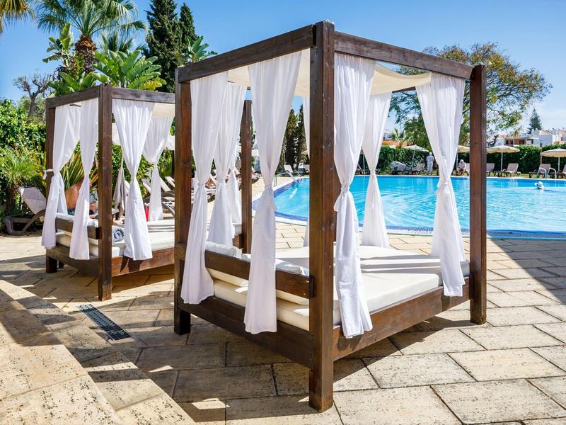 Holz-Überdachungen mit weißen Vorhängen stehen am Pool auf Steinfliesen unter blauem Himmel.
