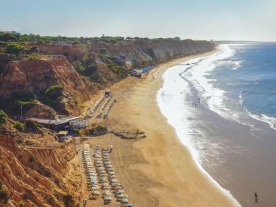 Langer Sandstrand mit Klippen und Liegestühlen unter blauem Himmel.