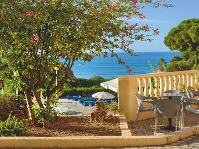 Terrasse mit Gartenmöbeln, blühendem Baum, Pool und Meer im Hintergrund an sonnigem Tag.