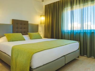 Modernes Schlafzimmer mit großem Bett, grünen Kissen, Decke und Vorhängen bei Tageslicht.