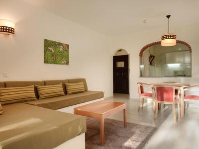 Helles Wohnzimmer mit braunem Ecksofa, Holztisch, Teppich und Esstisch mit roten Stühlen.