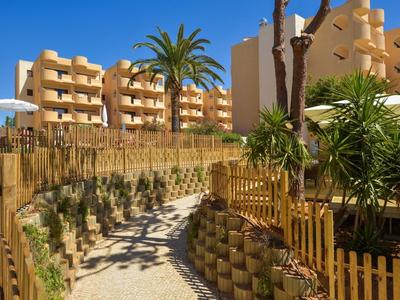 Sonniger Weg mit Palmen, Bäumen und runden Pflanztöpfen vor modernen beigen Gebäuden.