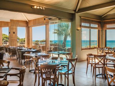 Helles Restaurant mit Holztischen und Stühlen, großen Fenstern und Blick auf das Meer.
