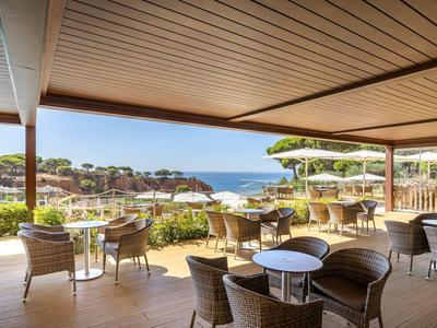 Überdachte Terrasse mit Holzdielen, runden Tischen und Korbsesseln, Blick aufs Meer und grüne Bäume.
