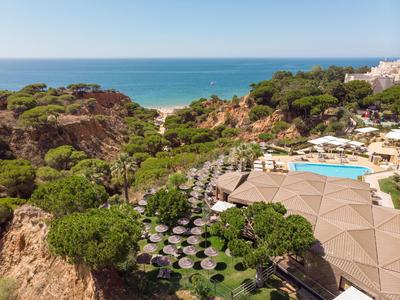 Landschaft mit Felsen, grünen Bäumen, Pool, Sonnenschirmen und Blick auf das blaue Meer.