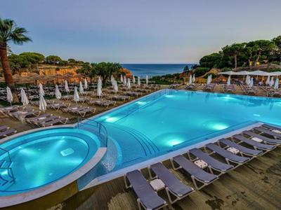 Großer beleuchteter Pool mit Liegestühlen, Sonnenschirmen und Palmen, Meer im Hintergrund.