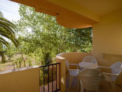 Balkon mit weißen Plastikstühlen und Tisch, umgeben von grünen Bäumen und gelben Wänden.