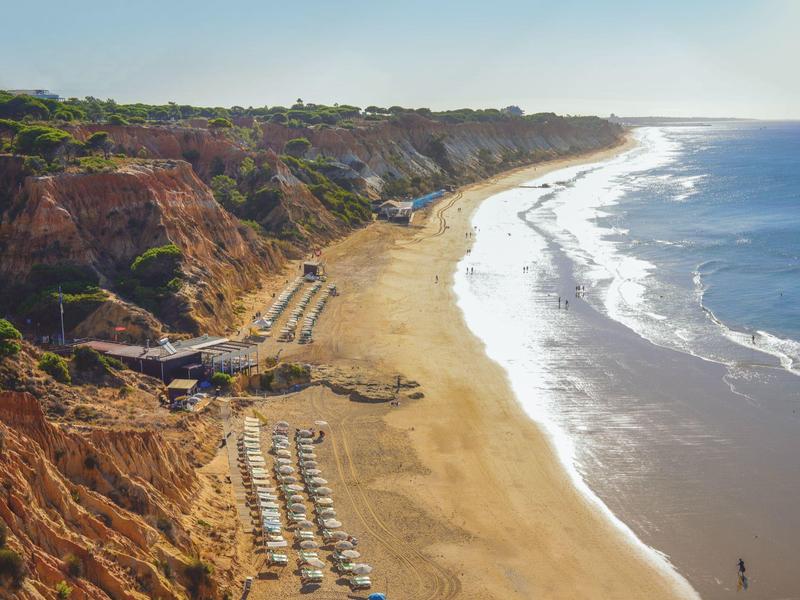 Langer Sandstrand mit Klippen und Liegestühlen unter blauem Himmel.