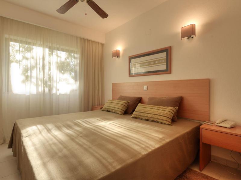 Helles Schlafzimmer mit Doppelbett, Holzkopfteil, zwei Kissen, Nachttisch und Deckenventilator.