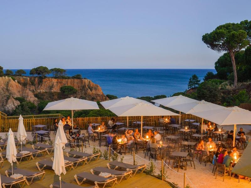 Strandrestaurant mit weißen Sonnenschirmen, Holzmöbeln und Blick auf Felsen und Meer bei Abendlicht.