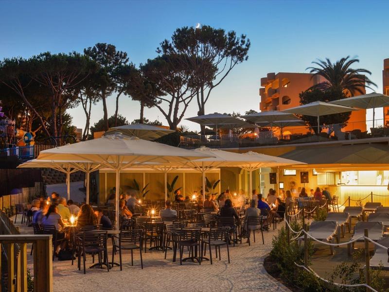 Abendliche Szene mit Gästen in einem offenen Restaurantbereich unter Bäumen und beleuchteten Gebäuden.