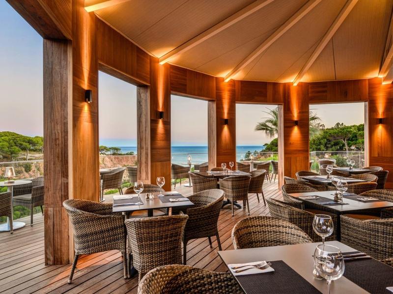 Offenes Restaurant mit Holzmöbeln und Blick auf das Meer, Palmen und einen hellen Himmel.