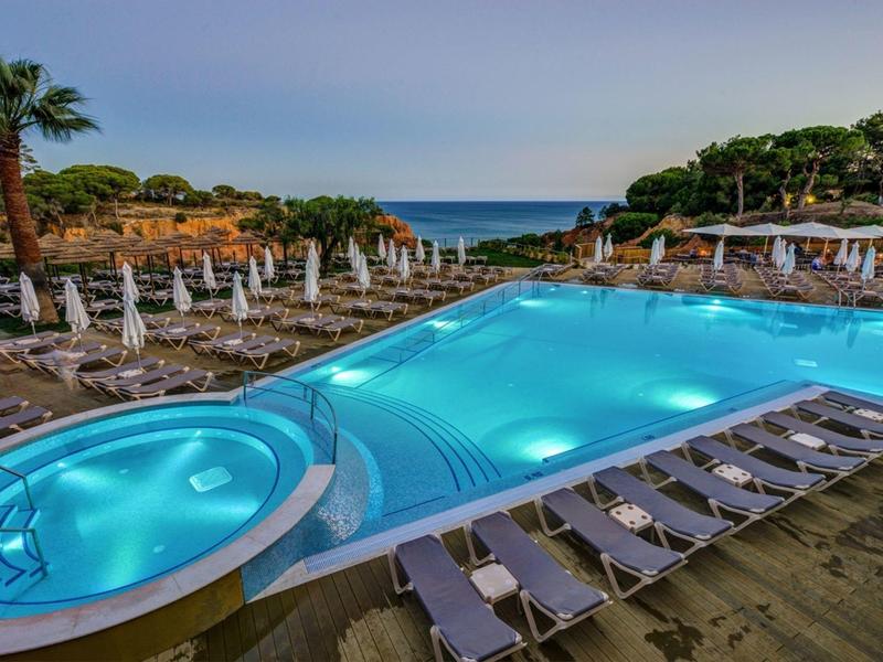 Großer beleuchteter Pool mit Liegestühlen, Sonnenschirmen und Palmen, Meer im Hintergrund.