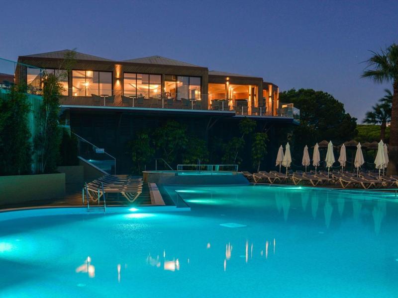 Beleuchteter Pool in der Dämmerung vor einem Hotel mit Terrasse und Palmen.