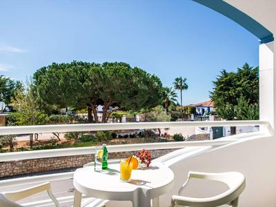 Balkon mit weißem Tisch und Stühlen, Getränke und Blumen, grüner Garten im Hintergrund, blauer Himmel.