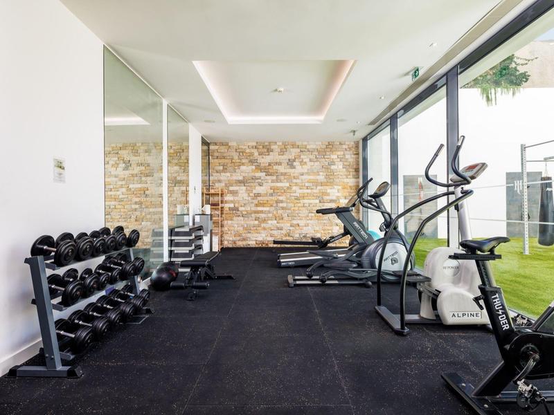 Moderní posilovna s činkami, běžeckými pásy a eliptickými trenažéry u velkých oken.