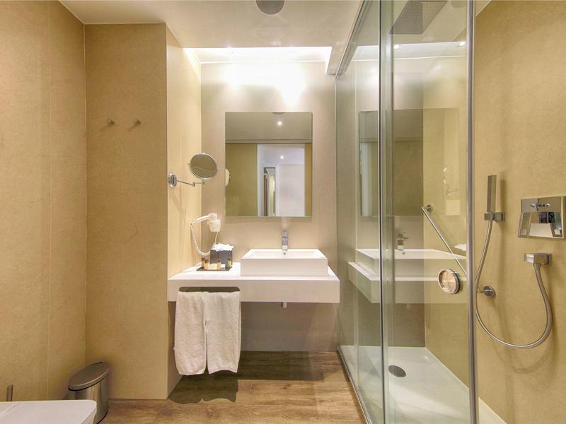 Moderní hotelová koupelna se skleněnou sprchou, umyvadlem a bílými ručníky.