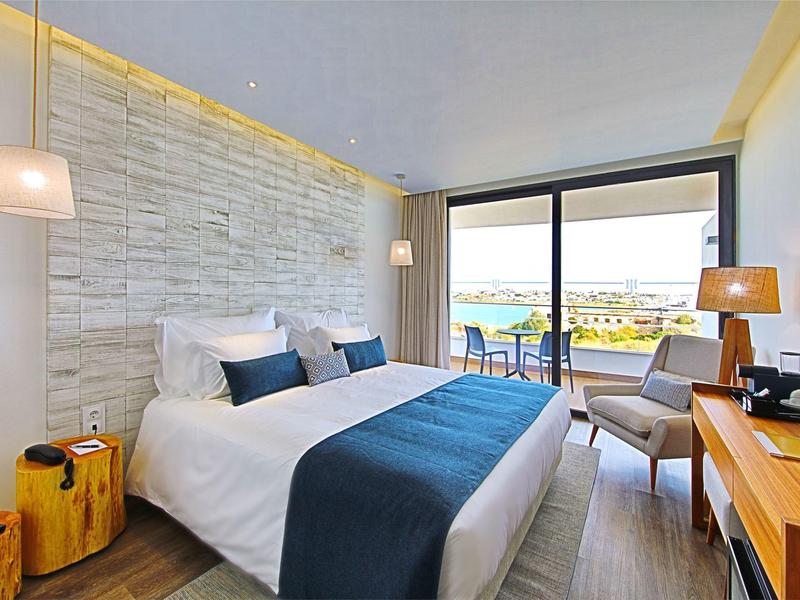 Světly hotelový pokoj s manželskou postelí, křeslem, psacím stolem a balkónem s výhledem na moře.