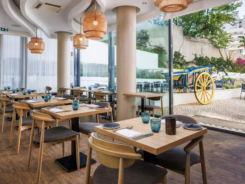 Moderní restaurace s dřevěným nábytkem a velkými okny s výhledem do zahrady.