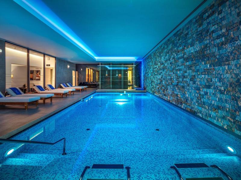Elegantní krytý bazén s modrým osvětlením a lehátky v moderní hotelové části