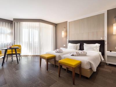 Chambre d'hôtel moderne avec lit double, tabourets jaunes et grande fenêtre avec rideaux.