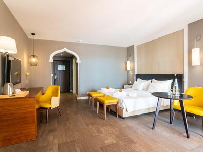 Chambre d'hôtel lumineuse avec lit double, fauteuils jaunes, bureau et sol en bois foncé.