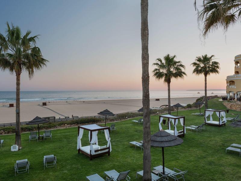 Coucher de soleil à la plage avec des palmiers, des transats et des petites cabanes sur une pelouse verte.