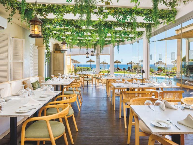 Salle de restaurant lumineuse et moderne avec chaises en bois, tables dressées et vue sur la mer.
