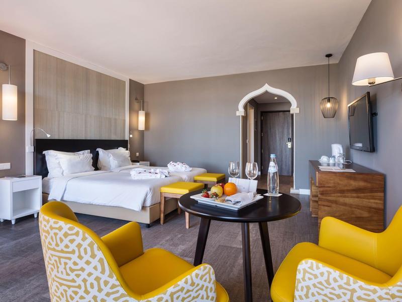 Chambre d'hôtel lumineuse avec deux lits, fauteuils jaunes, sol en bois et table ronde avec des fruits.